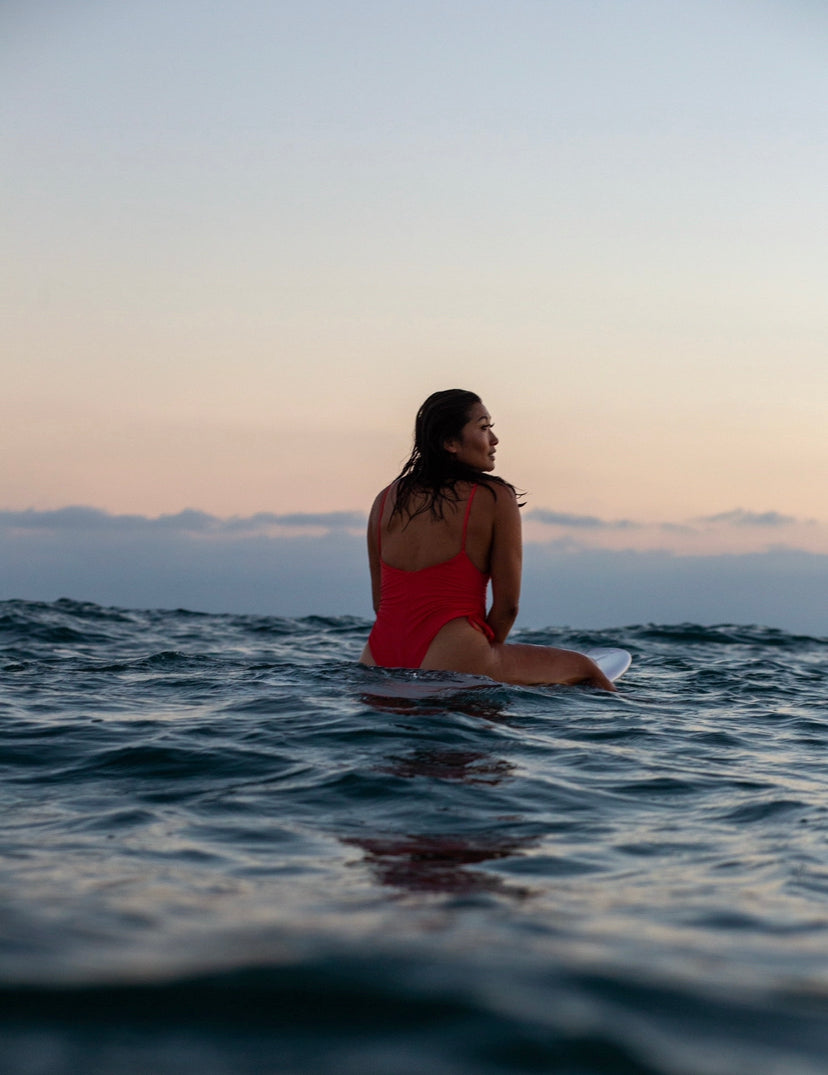 A person in a cheeky one piece swimsuit sits on a surfboard in the ocean at sunset, gazing toward the horizon with calm water and clouds in the distance.